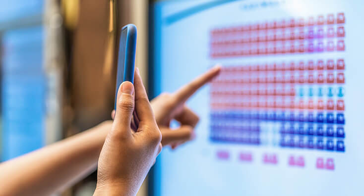 Person Pointing at a Computer Screen and Holding a Phone