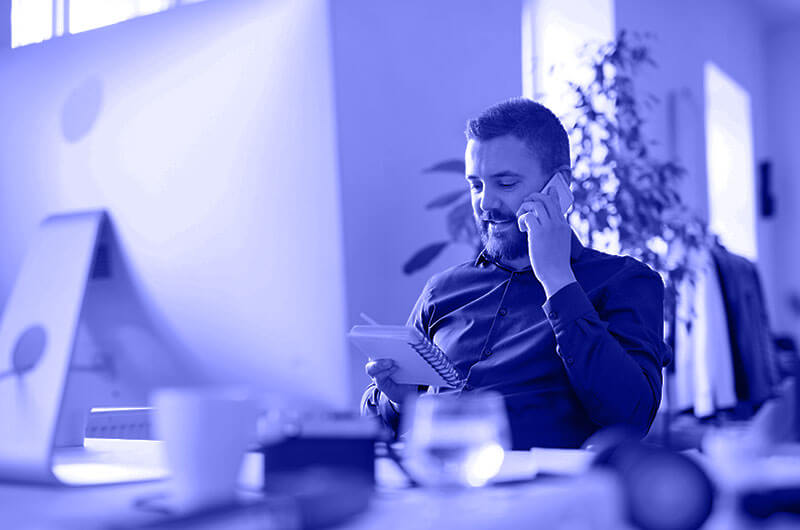 Blue Image of a Man Sat Down at a Desk on the Phone and Looking at a Notebook