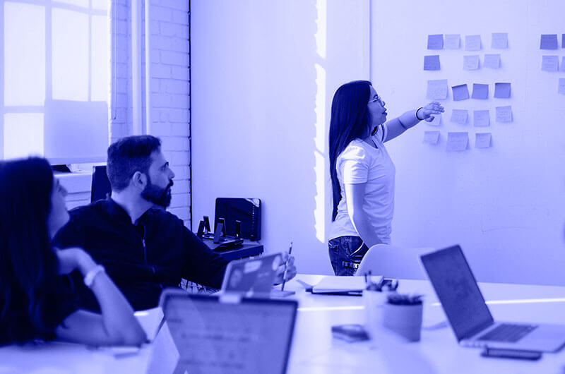 Blue Image of a Meeting where a Woman is Showing People Post It Notes on a Whiteboard