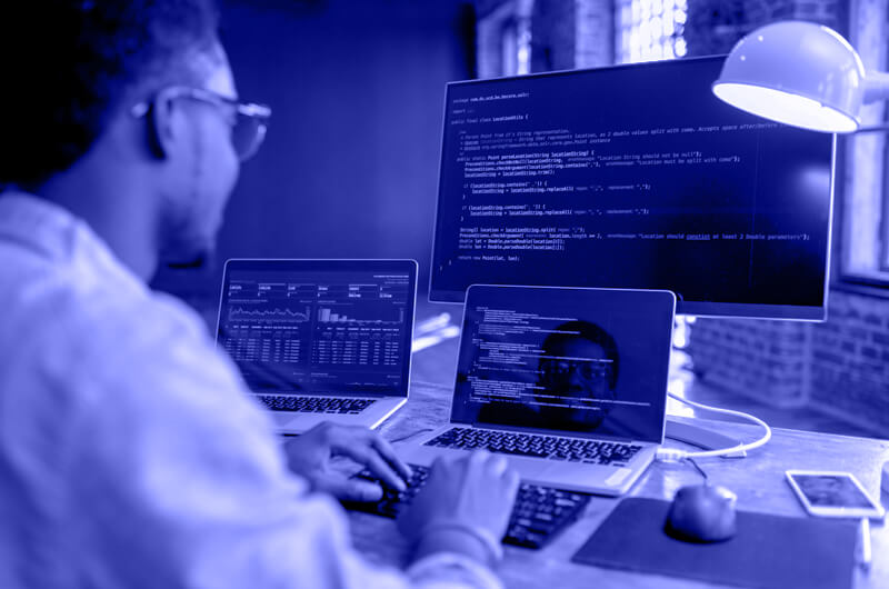 Blue Image of a Man Working on a Desktop and Two Laptops