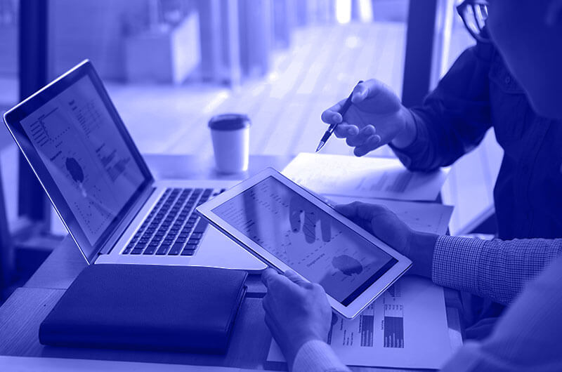 Blue Image of Two People Discussing Graphs On a Tablet Laptop and Paper Documents