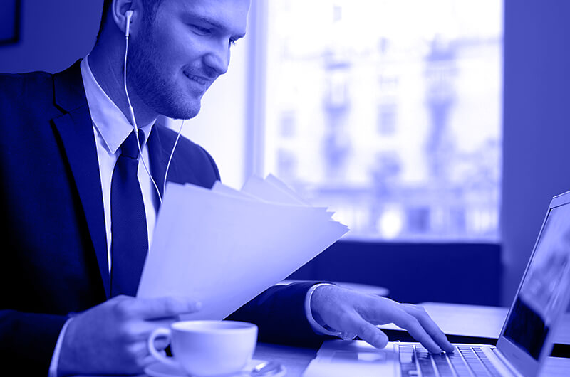 Blue Image of a Man Working on a Laptop Holding Paper Documents and Wearing Earphones at a Table