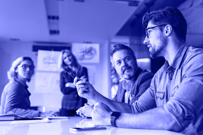 Blue Image of a Man Talking to Three Other People in a Meeting sat at a Table